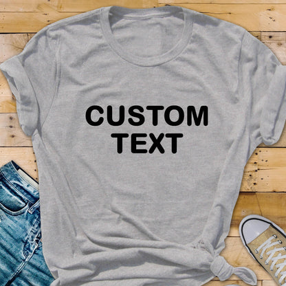 May include: A light gray short-sleeved t-shirt featuring the words CUSTOM TEXT printed in bold black capital letters across the center of the chest. The shirt has a crew neckline and is made of a soft, heathered fabric with a casual fit.