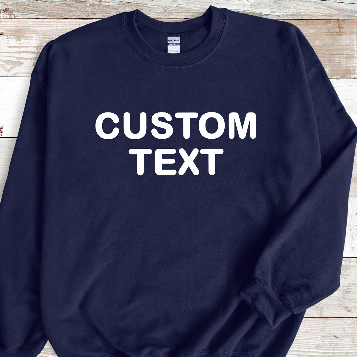 May include: A navy blue crewneck sweatshirt featuring the words CUSTOM TEXT printed in bold white capital letters across the chest. The garment has long sleeves, a ribbed collar, and a white brand label visible at the inside of the neckline.