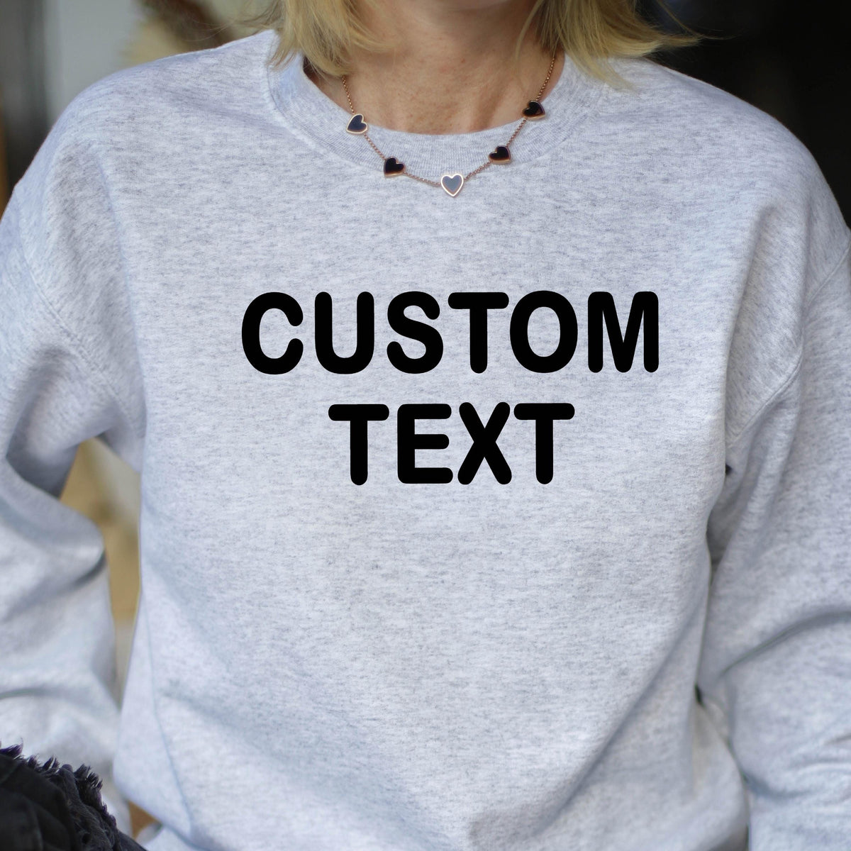 May include: A light gray heather crewneck sweatshirt featuring the words CUSTOM TEXT printed in bold black capital letters across the chest. The garment has long sleeves and a ribbed neckline, providing a simple and functional design for casual wear.