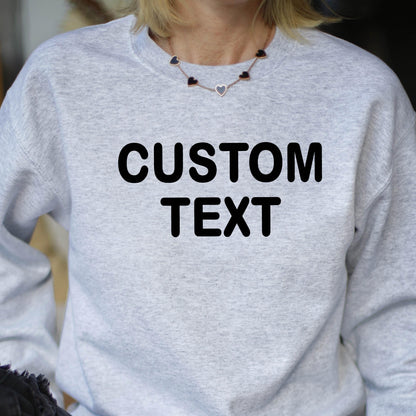 May include: A light gray heather crewneck sweatshirt featuring the words CUSTOM TEXT printed in bold black capital letters across the chest. The garment has long sleeves and a ribbed neckline, providing a simple and functional design for casual wear.