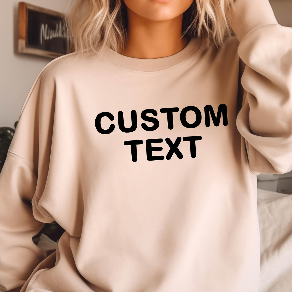 May include: A beige crewneck sweatshirt featuring the words CUSTOM TEXT printed in bold black capital letters across the chest. The garment has long sleeves and a relaxed fit, providing a neutral base for the prominent dark text design.