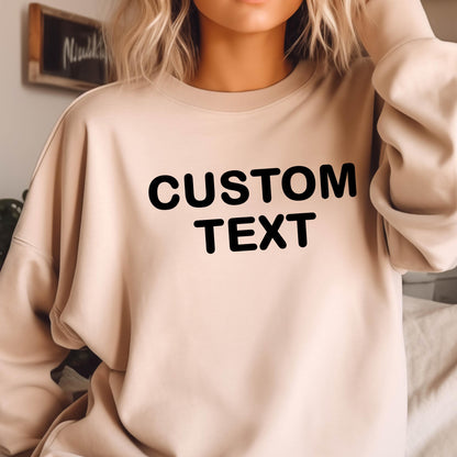 May include: A beige crewneck sweatshirt featuring the words CUSTOM TEXT printed in bold black capital letters across the chest. The garment has long sleeves and a relaxed fit, providing a neutral base for the prominent dark text design.