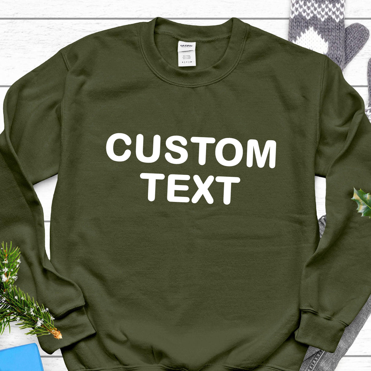 May include: An olive green long-sleeve crewneck sweatshirt featuring the words CUSTOM TEXT printed in large white capital letters across the chest. The garment has a ribbed neckline, cuffs, and hem, with a white brand label inside the collar.