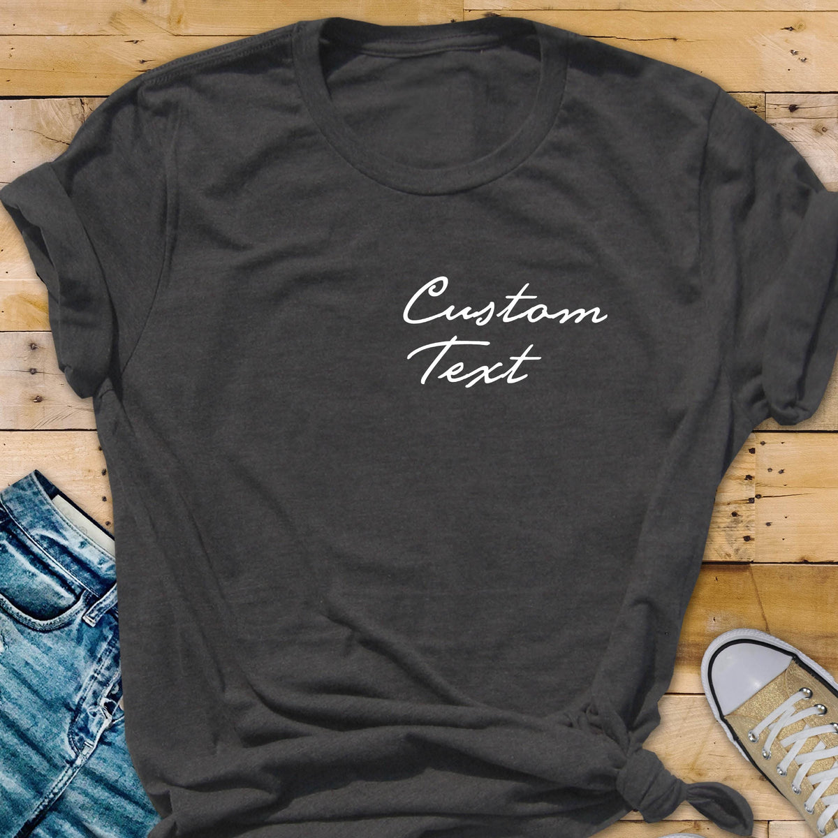 May include: Dark gray t-shirt with the words "Custom Text" in white script. The shirt is knotted at the bottom and has rolled-up sleeves. The background includes a pair of blue jeans and a canvas shoe.