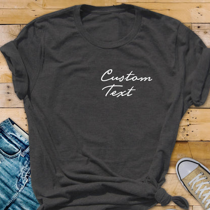 May include: Dark gray t-shirt with the words "Custom Text" in white script. The shirt is knotted at the bottom and has rolled-up sleeves. The background includes a pair of blue jeans and a canvas shoe.