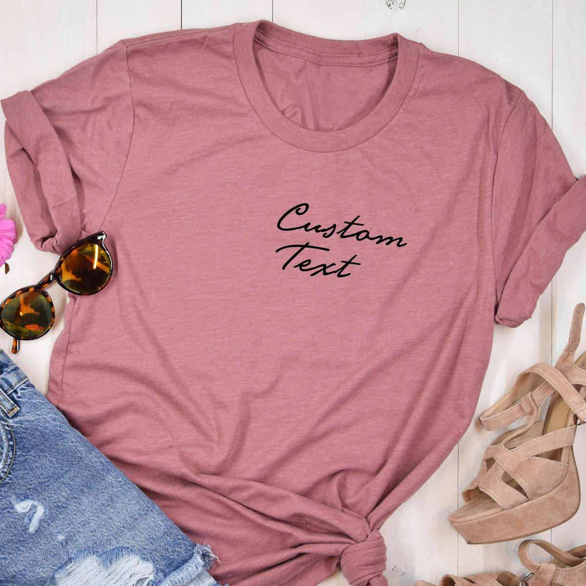 May include: A dusty rose t-shirt with the words "Custom Text" in black script. The shirt is paired with sunglasses, denim shorts, and platform sandals. The shirt is knotted at the bottom.