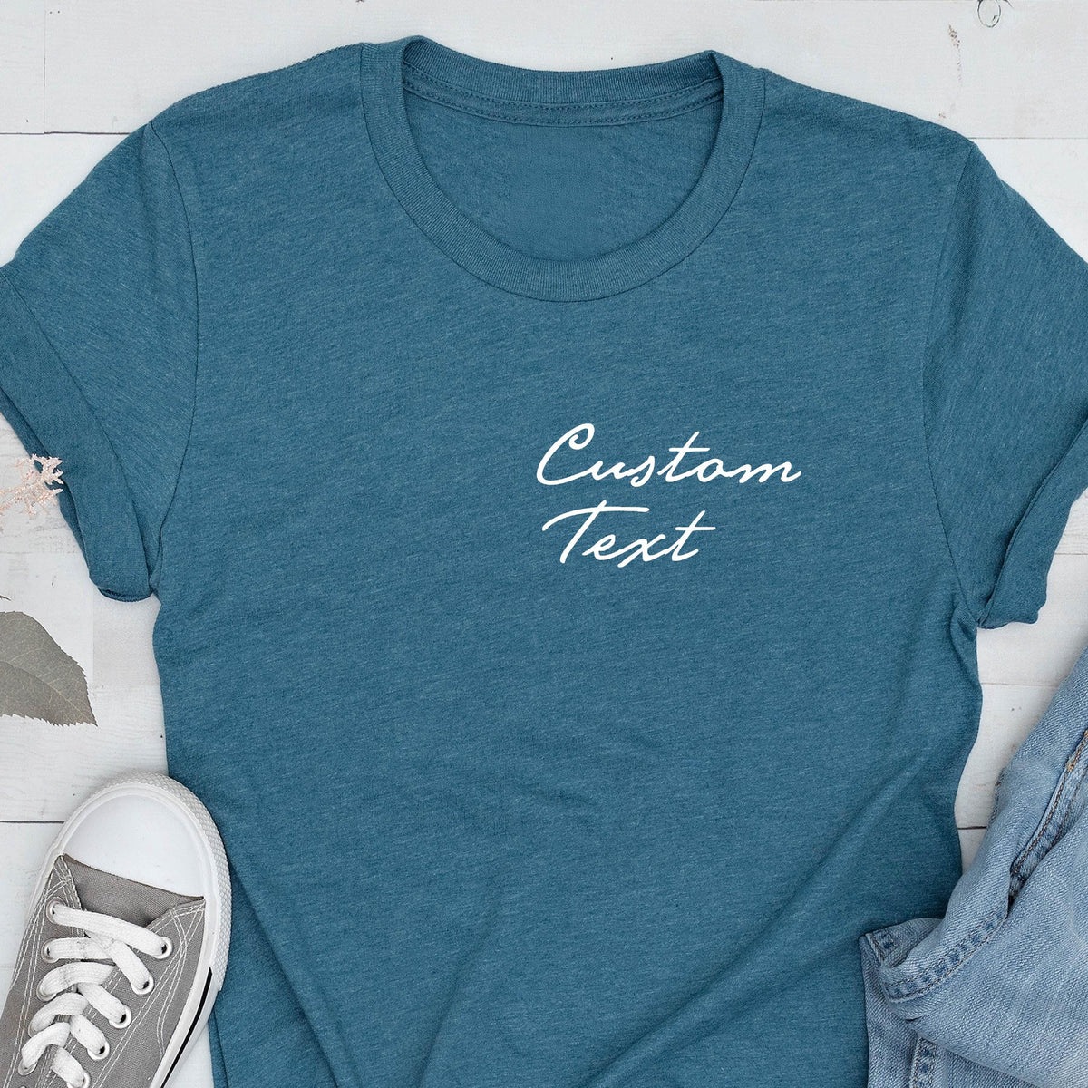 May include: Teal-colored t-shirt with the words "Custom Text" in white script. The shirt has short sleeves and a crew neck. A pair of gray sneakers is visible in the lower left corner.