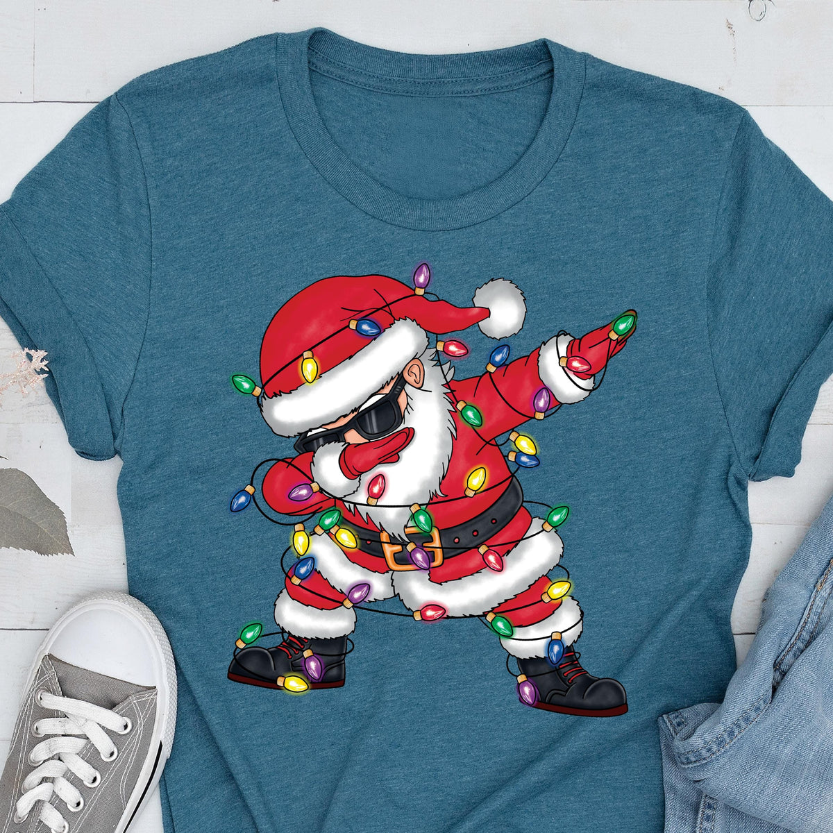 May include: Teal t-shirt featuring a cartoon Santa Claus dabbing. Santa is wearing sunglasses, a red suit, and a hat, and is wrapped in Christmas lights. The design is a fun, festive take on a holiday theme.