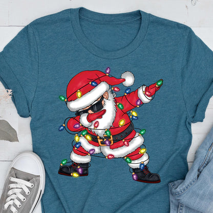 May include: Teal t-shirt featuring a cartoon Santa Claus dabbing. Santa is wearing sunglasses, a red suit, and a hat, and is wrapped in Christmas lights. The design is a fun, festive take on a holiday theme.