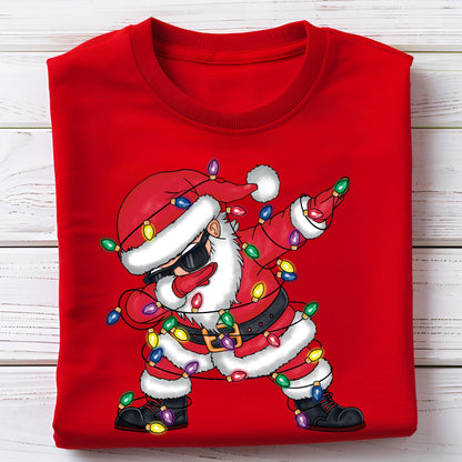 May include: Red sweatshirt featuring a cartoon Santa Claus dabbing. Santa wears sunglasses, a red hat, and a black belt. He is decorated with colorful Christmas lights. The sweatshirt is folded on a white wooden surface.