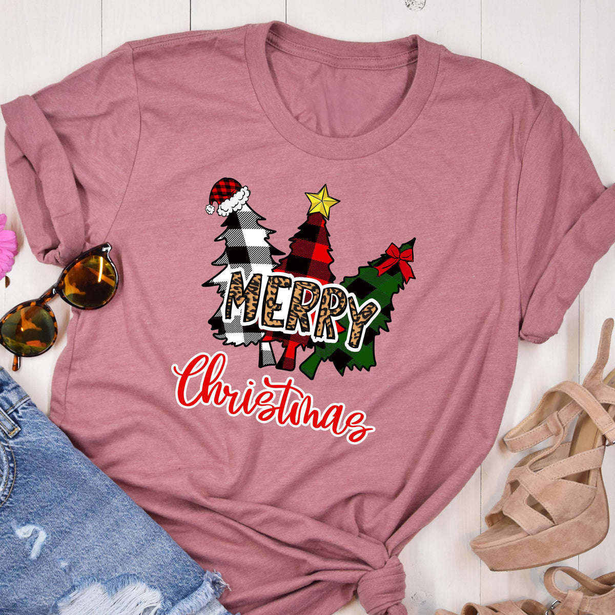 May include: A mauve short-sleeved t-shirt featuring a graphic of three stylized Christmas trees in black and white buffalo plaid, red and black plaid, and green plaid. The word MERRY is in leopard print, and Christmas is in red script.