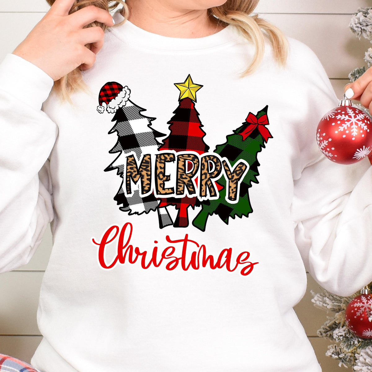 May include: A white long-sleeve sweatshirt featuring a graphic of three stylized Christmas trees in black and white buffalo plaid, red and black buffalo plaid, and green and black buffalo plaid. The word MERRY is in leopard print.