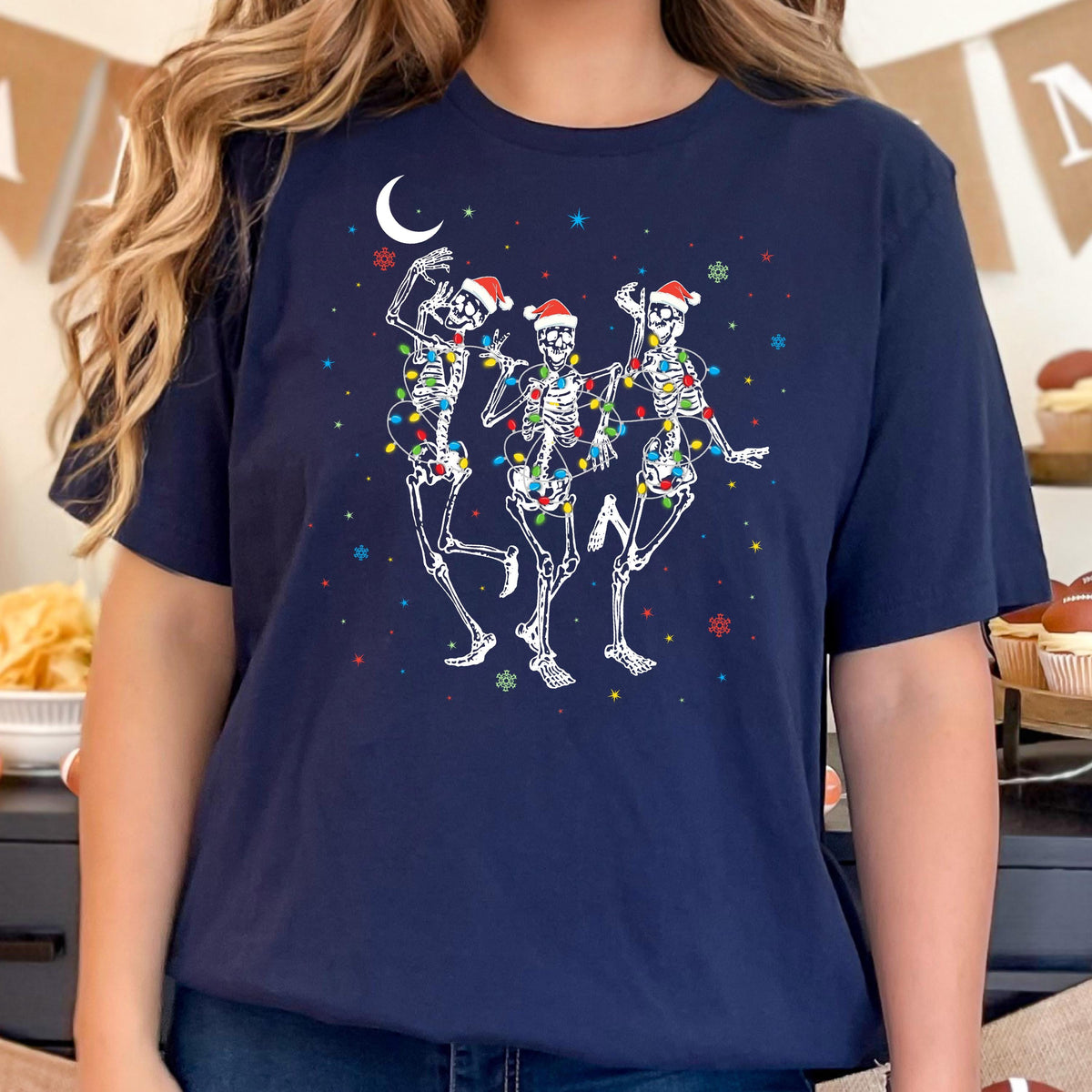 May include: Navy blue t-shirt featuring a festive design of three dancing skeletons adorned with Santa hats and colorful Christmas lights. The skeletons are set against a backdrop of a crescent moon and scattered stars, creating a whimsical holiday theme.