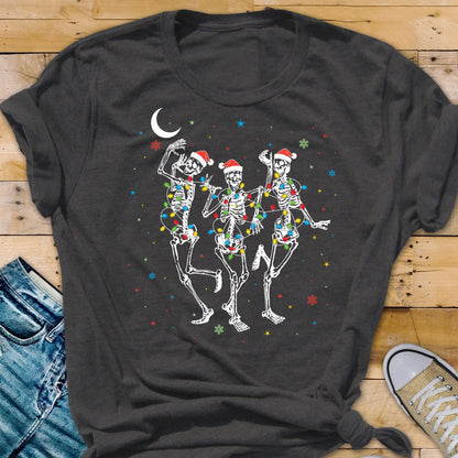 May include: Dark gray t-shirt featuring a festive design of three dancing skeletons. Each skeleton wears a Santa hat and is adorned with colorful Christmas lights. A crescent moon and snowflakes complete the holiday theme.