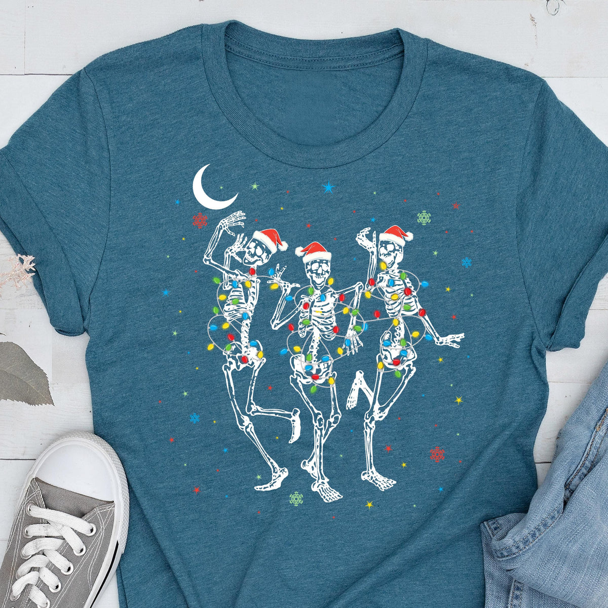 May include: Teal t-shirt featuring a festive design of three dancing skeletons. Each skeleton wears a Santa hat and is adorned with colorful Christmas lights. A crescent moon and snowflakes complete the holiday-themed graphic.