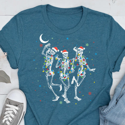 May include: Teal t-shirt featuring a festive design of three dancing skeletons. Each skeleton wears a Santa hat and is adorned with colorful Christmas lights. A crescent moon and snowflakes complete the holiday-themed graphic.