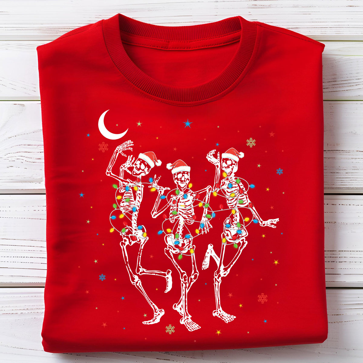 May include: A red sweatshirt featuring a festive design of three dancing skeletons wearing Santa hats and wrapped in colorful Christmas lights. The design includes a crescent moon, stars, and snowflakes, creating a holiday theme.