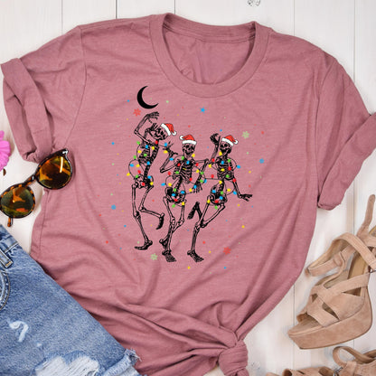 May include: A dusty rose t-shirt featuring a whimsical illustration of three dancing skeletons adorned with Santa hats and colorful string lights. A crescent moon and stars are also in the design. The shirt is folded, with sunglasses and denim shorts visible.