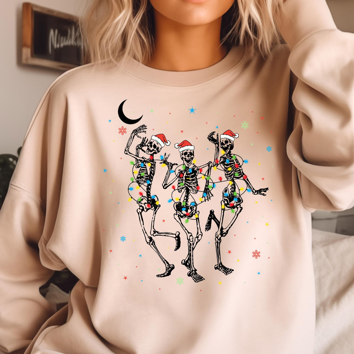 May include: A beige crewneck sweatshirt featuring a graphic print of three skeletons wearing red and white Santa hats. The skeletons are wrapped in colorful Christmas lights and are surrounded by small blue stars, red snowflakes, and a black crescent moon.