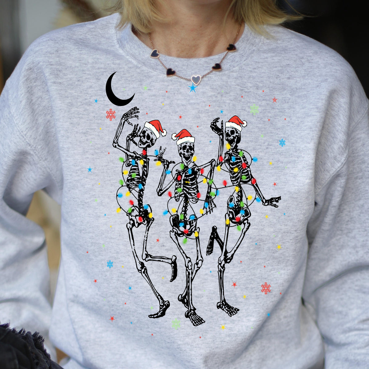 May include: A light gray crewneck sweatshirt featuring a graphic of three skeletons wearing red and white Santa hats. The skeletons are tangled in colorful Christmas lights, surrounded by small red and blue stars and snowflakes, with a black crescent moon.