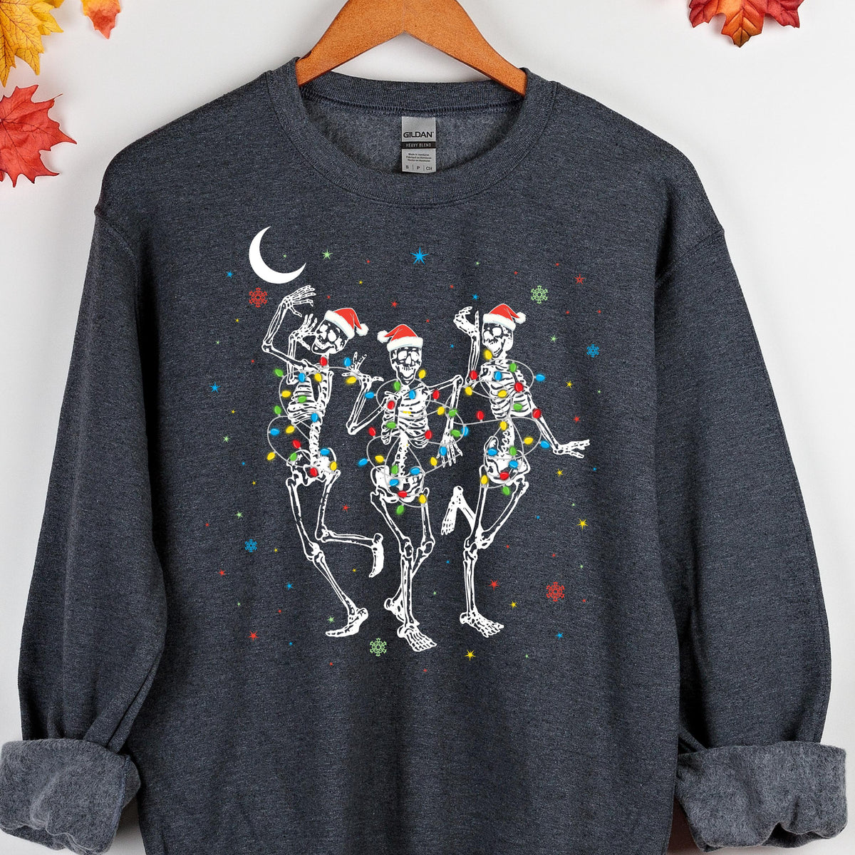 May include: A dark heather gray crewneck sweatshirt featuring a graphic of three white skeletons wearing red Santa hats. The skeletons are wrapped in colorful Christmas light strings, surrounded by a white crescent moon and small snowflakes.