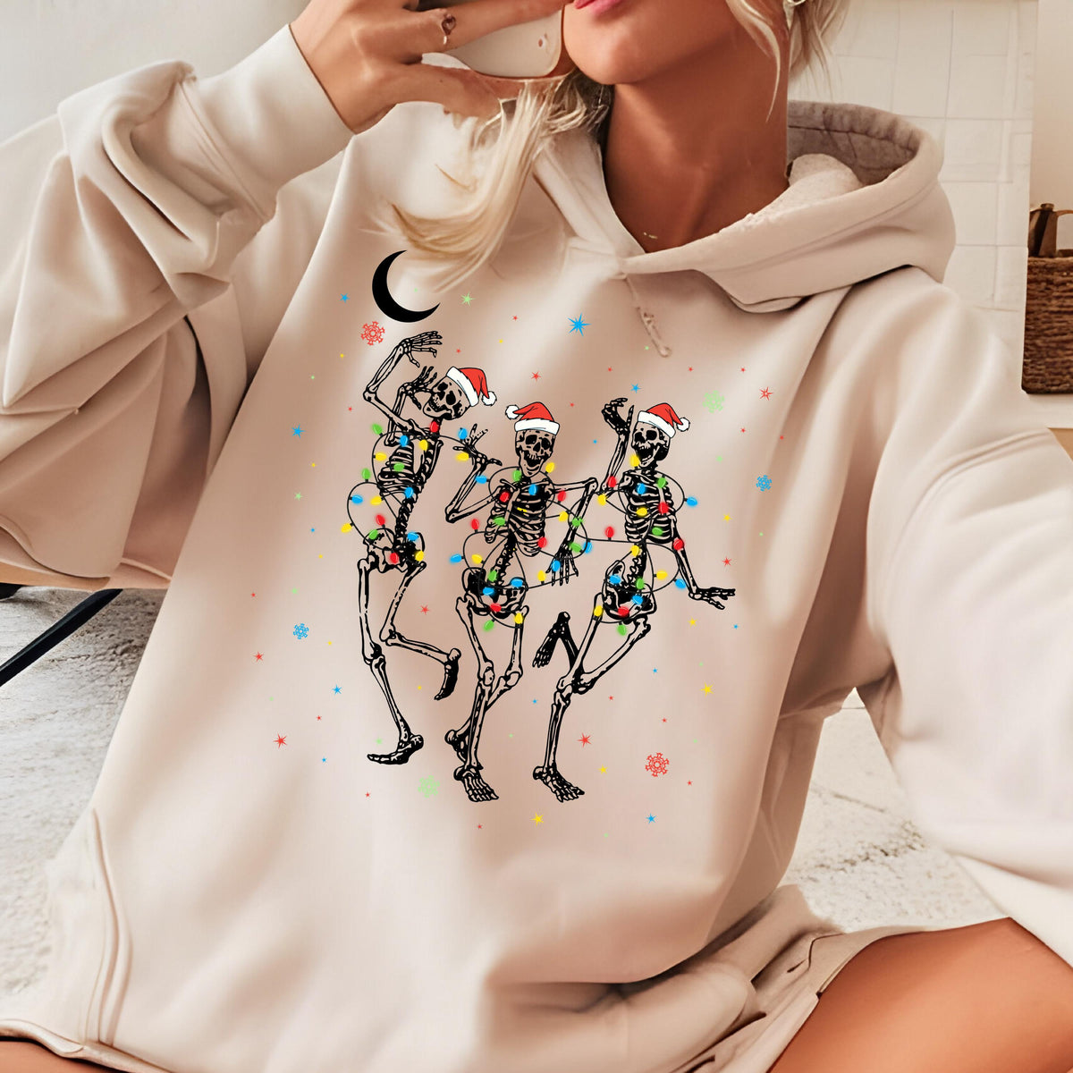 May include: A beige hooded sweatshirt featuring a graphic print of three skeletons wearing red and white Santa hats. The skeletons are tangled in colorful Christmas lights, surrounded by small stars, snowflakes, and a black crescent moon.