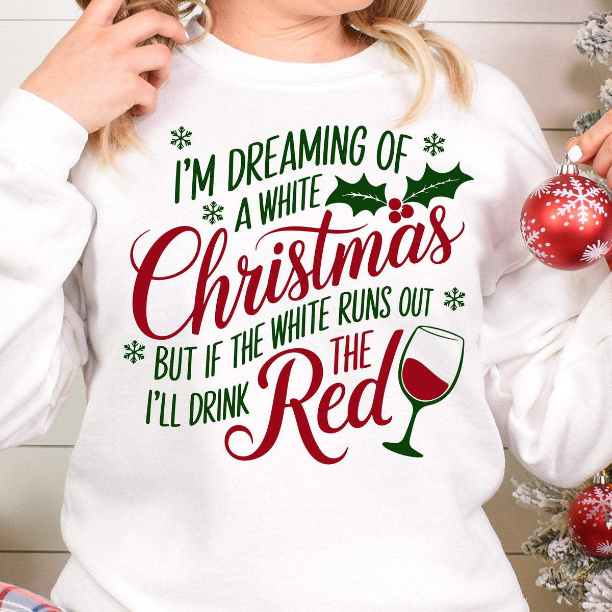 May include: A white long-sleeve sweatshirt featuring green and red text that reads I'M DREAMING OF A WHITE CHRISTMAS BUT IF THE WHITE RUNS OUT I'LL DRINK THE RED. The design includes green holly leaves, red berries, snowflakes, and a red wine glass.