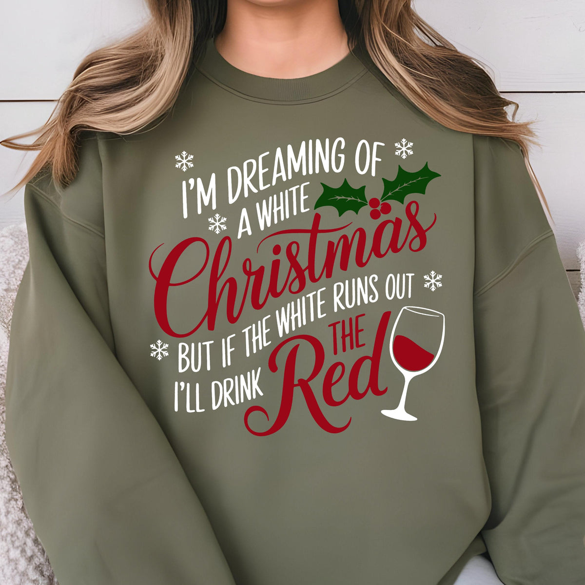 May include: An olive green crewneck sweatshirt featuring white and red text that reads I'M DREAMING OF A WHITE CHRISTMAS BUT IF THE WHITE RUNS OUT I'LL DRINK THE RED. The design includes white snowflakes, green holly leaves, red berries, and a wine glass.