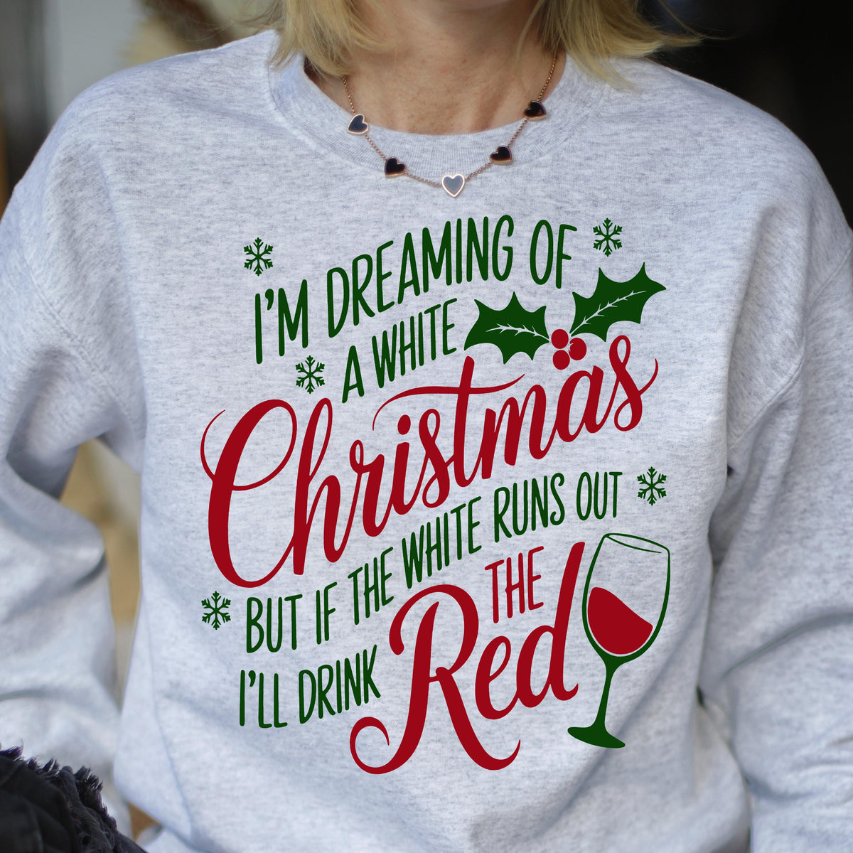 May include: A light gray crewneck sweatshirt featuring the green and red text I'M DREAMING OF A WHITE CHRISTMAS BUT IF THE WHITE RUNS OUT I'LL DRINK THE RED. The design includes green holly leaves, red berries, green snowflakes, and a wine glass.