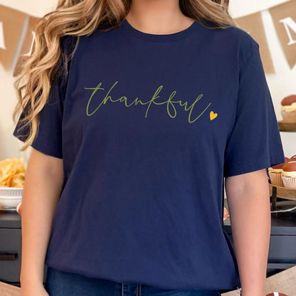 May include: Navy blue t-shirt with the word "Thankful" in a green script font and a small yellow heart. The shirt is a solid color and has a crew neck.