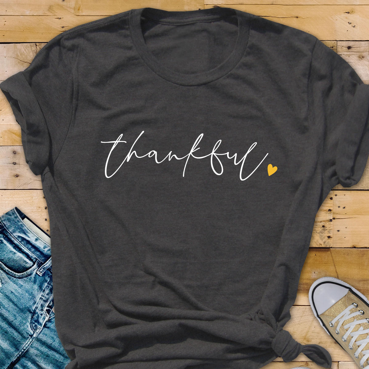 May include: Dark gray t-shirt with the word "thankful" in white script and a small yellow heart. The shirt is folded at the bottom and is paired with blue jeans and a pair of canvas shoes.