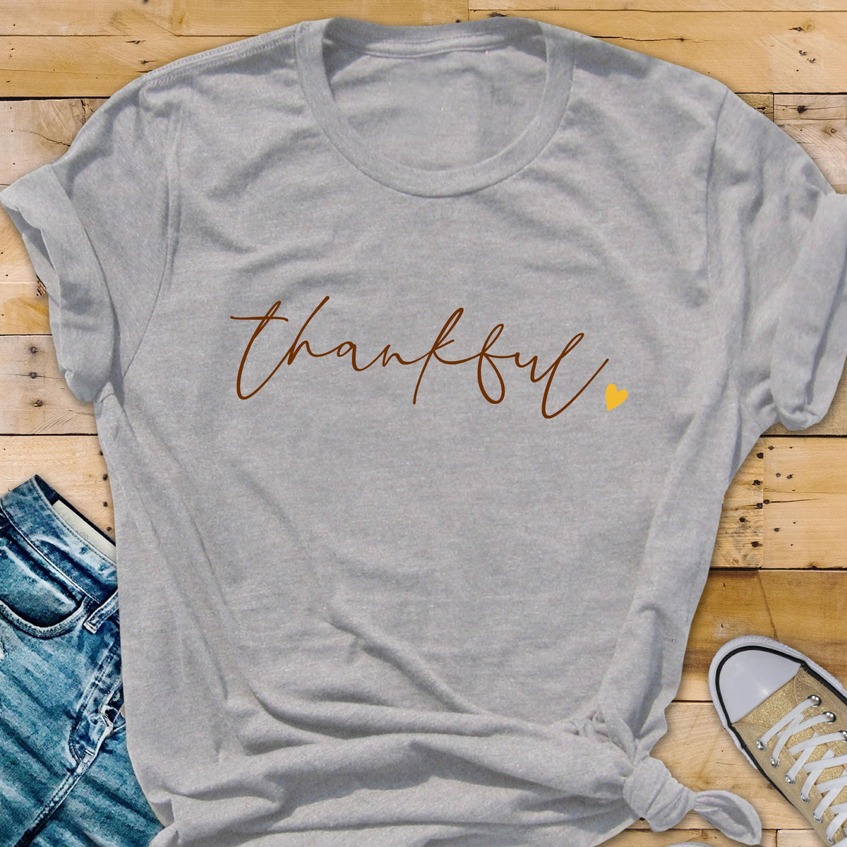 May include: A light gray t-shirt with the word "thankful" in a brown script font. A small yellow heart is next to the word. The shirt is knotted at the bottom. The background includes a pair of jeans and a canvas shoe.