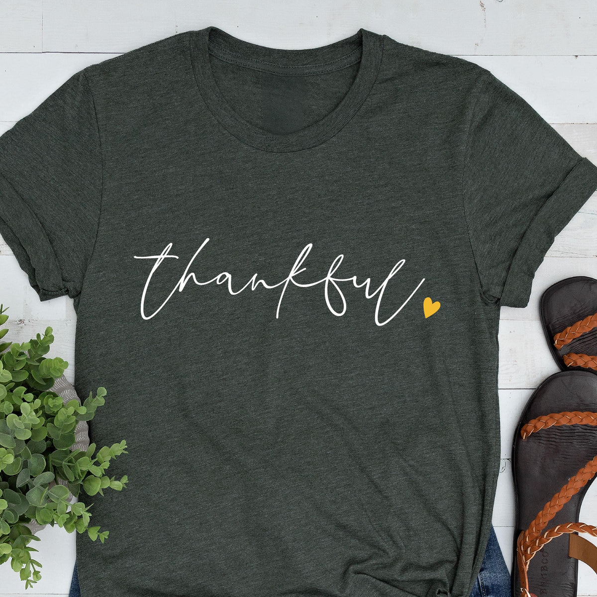 May include: Dark gray t-shirt with the word "thankful" in white script. A small yellow heart is next to the word. The shirt is folded, and part of a pair of sandals is visible in the corner.