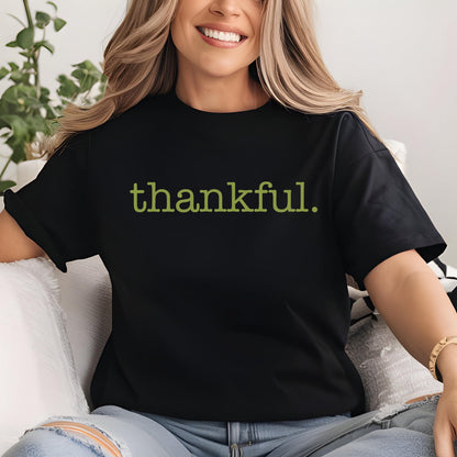 May include: Black t-shirt with the word "thankful" printed in a green, sans-serif font. The shirt is a solid black color and has short sleeves. The text is centered on the front of the shirt.