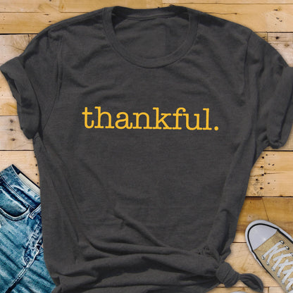 May include: Dark gray t-shirt with the word "thankful" printed in yellow. The shirt is made of a soft material and has a casual, relaxed fit. The text is in a simple, sans-serif font.