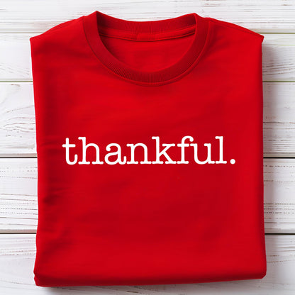 May include: A folded red sweatshirt with the word "thankful." printed in white, centered on the front. The sweatshirt has a crew neck and is made of a soft material. The background is a white wooden surface.