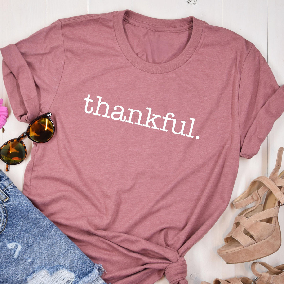 May include: A dusty rose t-shirt with the word "thankful" printed in white. The shirt is styled with denim shorts, sunglasses, and platform sandals. The t-shirt is folded at the sleeves.