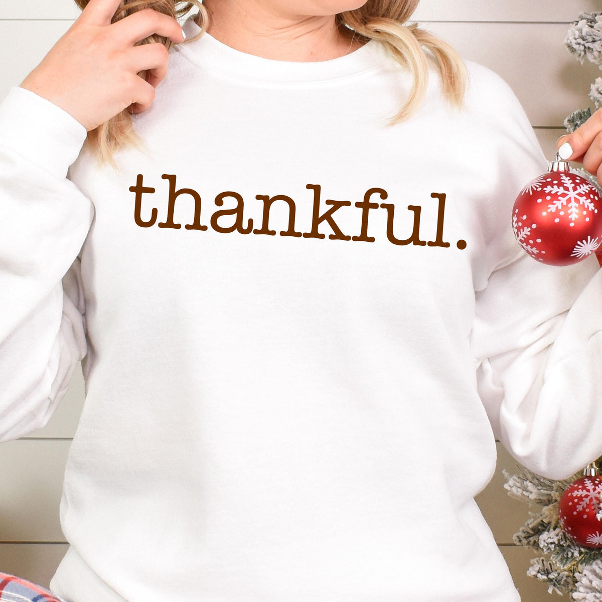 May include: White sweatshirt with the word "thankful" in brown lettering. The sweatshirt is a long-sleeved crew neck. A red and white Christmas ornament is held in the right hand. The overall theme is fall and Thanksgiving.