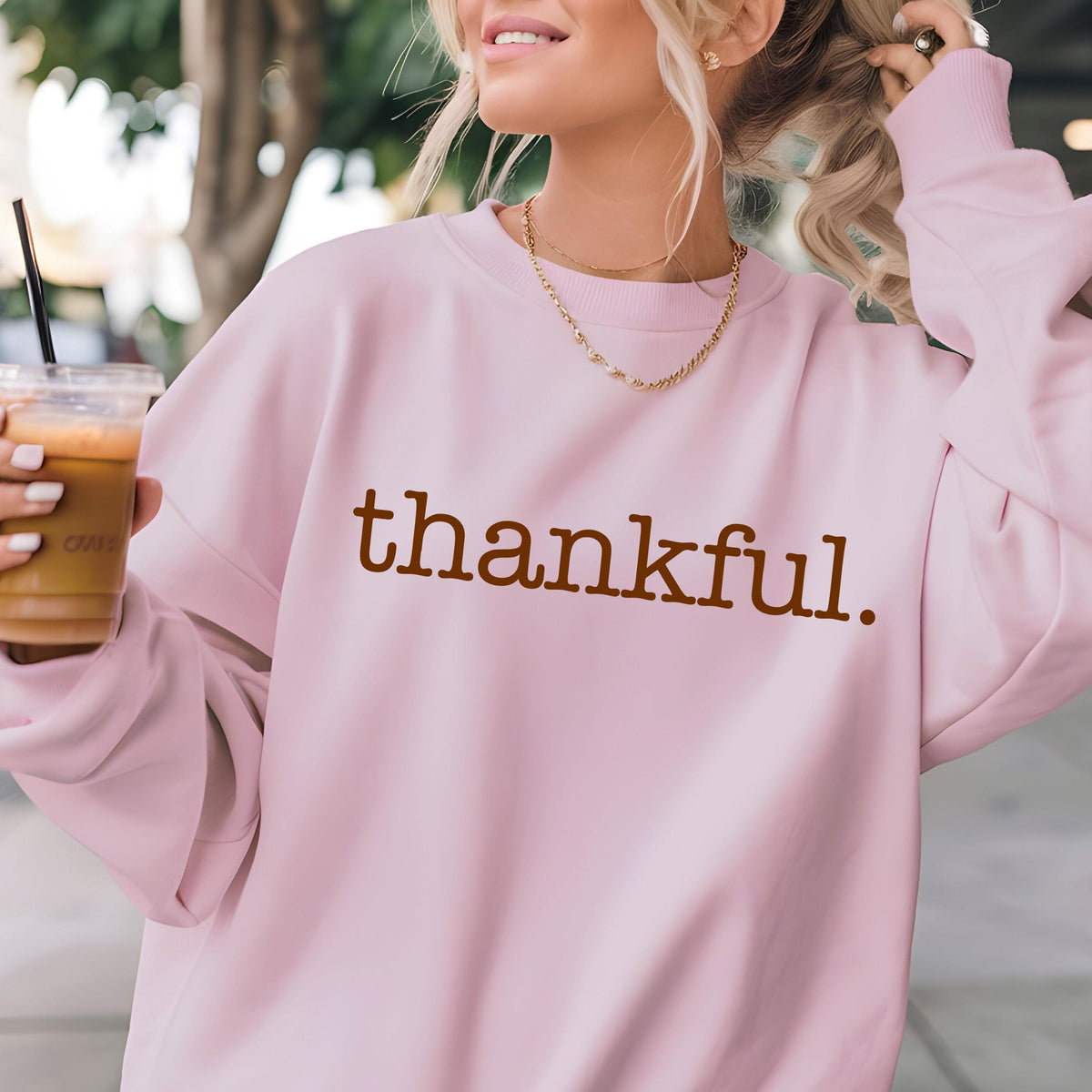 May include: A light pink sweatshirt with the word "thankful" printed in brown. The sweatshirt has long sleeves and a crew neck. A gold necklace is visible. The person is holding a clear plastic cup with a brown beverage and a black straw.