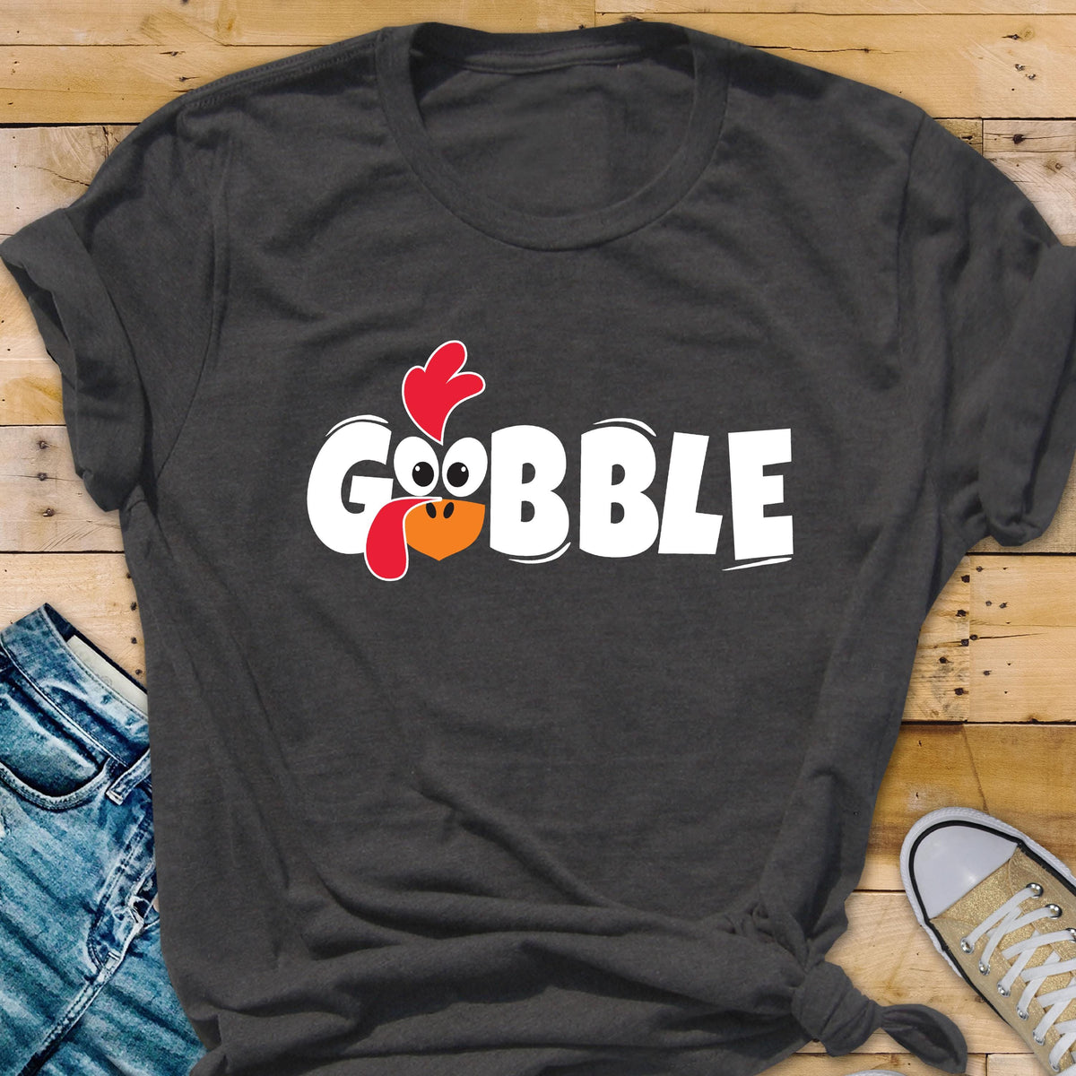 May include: A dark charcoal gray short-sleeve t-shirt featuring the white text GOBBLE printed across the chest. The letter O is replaced by a cartoon turkey face with a red wattle, a red comb, and an orange beak, all centered on the front.