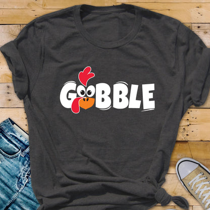 May include: A dark charcoal gray short-sleeve t-shirt featuring the white text GOBBLE printed across the chest. The letter O is replaced by a cartoon turkey face with a red wattle, a red comb, and an orange beak, all centered on the front.