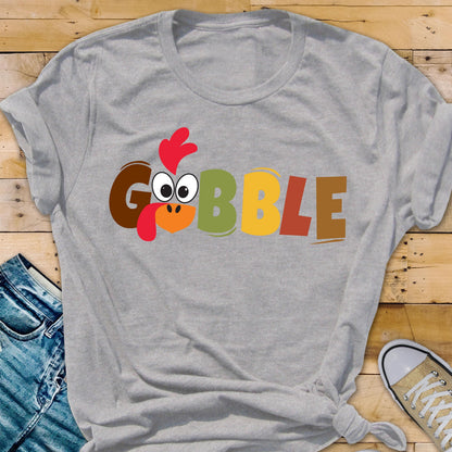 May include: A light gray short-sleeve t-shirt featuring the word GOBBLE printed in large, colorful block letters. The letter O is replaced by a cartoon turkey face with a red wattle, orange beak, and large white eyes with black pupils.