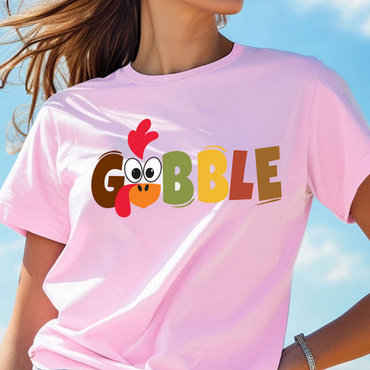 May include: A light pink short-sleeved t-shirt featuring the text GOBBLE in large, multi-colored block letters. The letter O is replaced by a cartoon turkey face with a red wattle, a yellow beak, and large white eyes with black pupils.