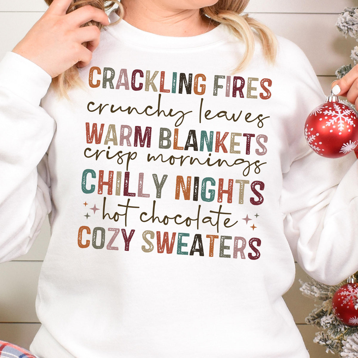 May include: White sweatshirt with a colorful graphic print that reads "Crackling Fires, Crunchy Leaves, Warm Blankets, Crisp Mornings, Chilly Nights, Hot Chocolate, Cozy Sweaters".