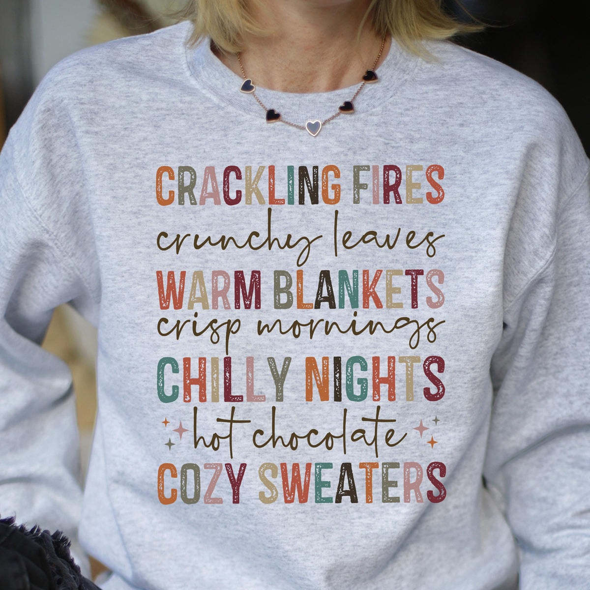 May include: Gray sweatshirt with a colorful text design that reads "Crackling Fires, Crunchy Leaves, Warm Blankets, Crisp Mornings, Chilly Nights, Hot Chocolate, Cozy Sweaters".