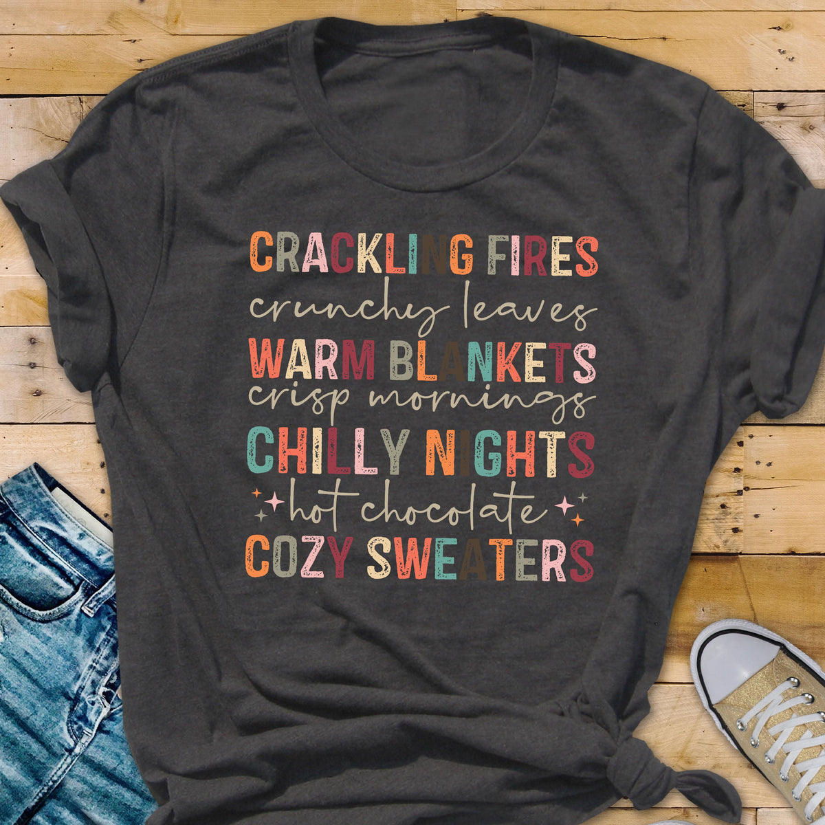 May include: Dark gray t-shirt with colorful text: "CRACKLING FIRES, crunchy leaves, WARM BLANKETS, crisp mornings, CHILLY NIGHTS, hot chocolate, COZY SWEATERS." The text is in various fonts and colors, evoking a cozy autumn theme.