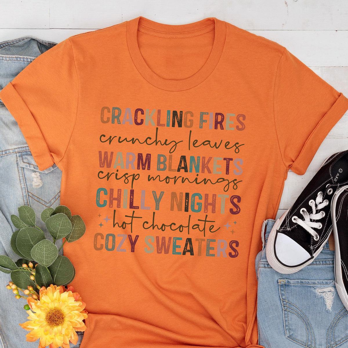 May include: Orange t-shirt with the text "CRACKLING FIRES, crunchy leaves, WARM BLANKETS, crisp mornings, CHILLY NIGHTS, hot chocolate, COZY SWEATERS" in various colors. The shirt is folded at the sleeves.