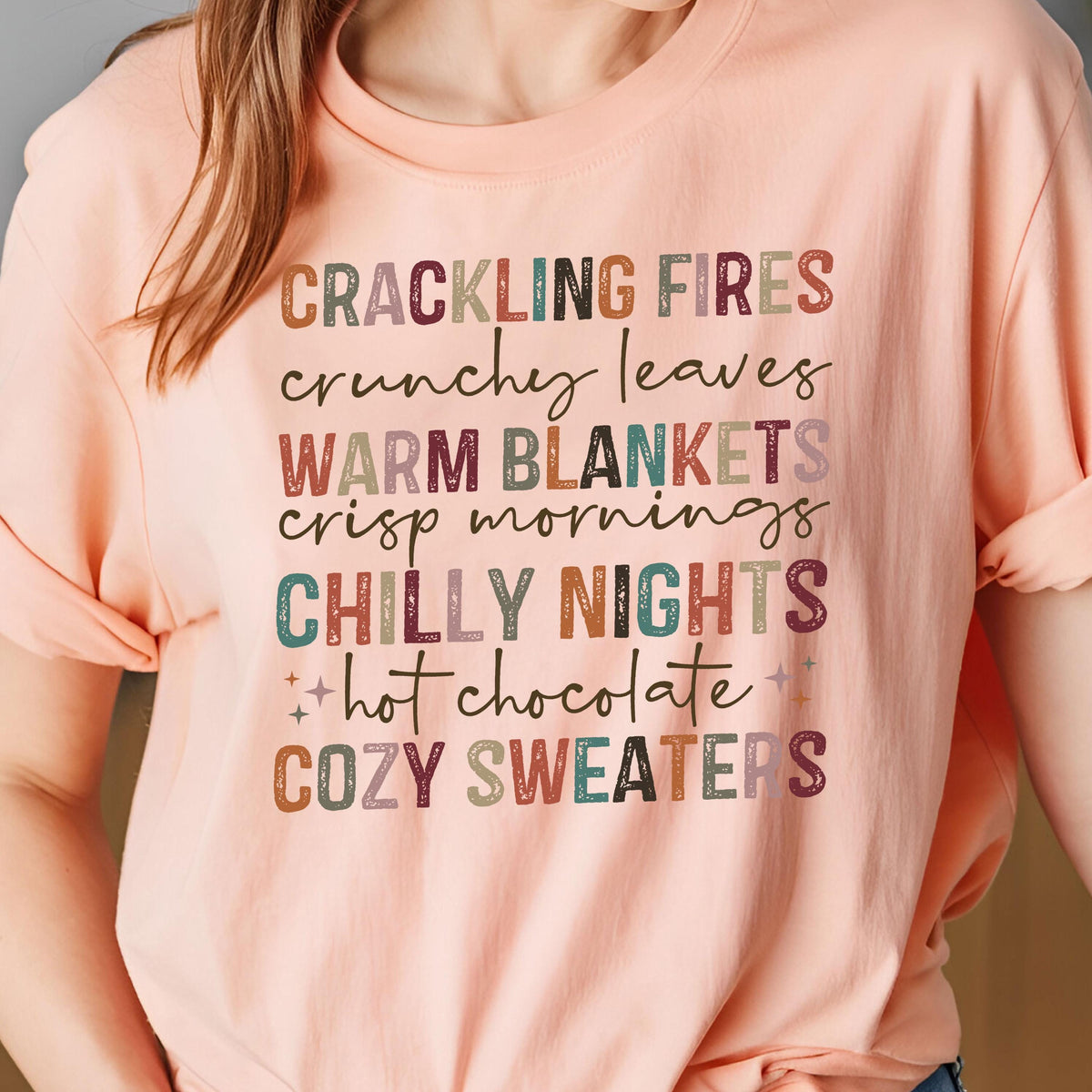 May include: A peach-colored t-shirt with the words "CRACKLING FIRES, crunchy leaves, WARM BLANKETS, crisp mornings, CHILLY NIGHTS, hot chocolate, COZY SWEATERS" in a variety of fall colors. The shirt has short sleeves.