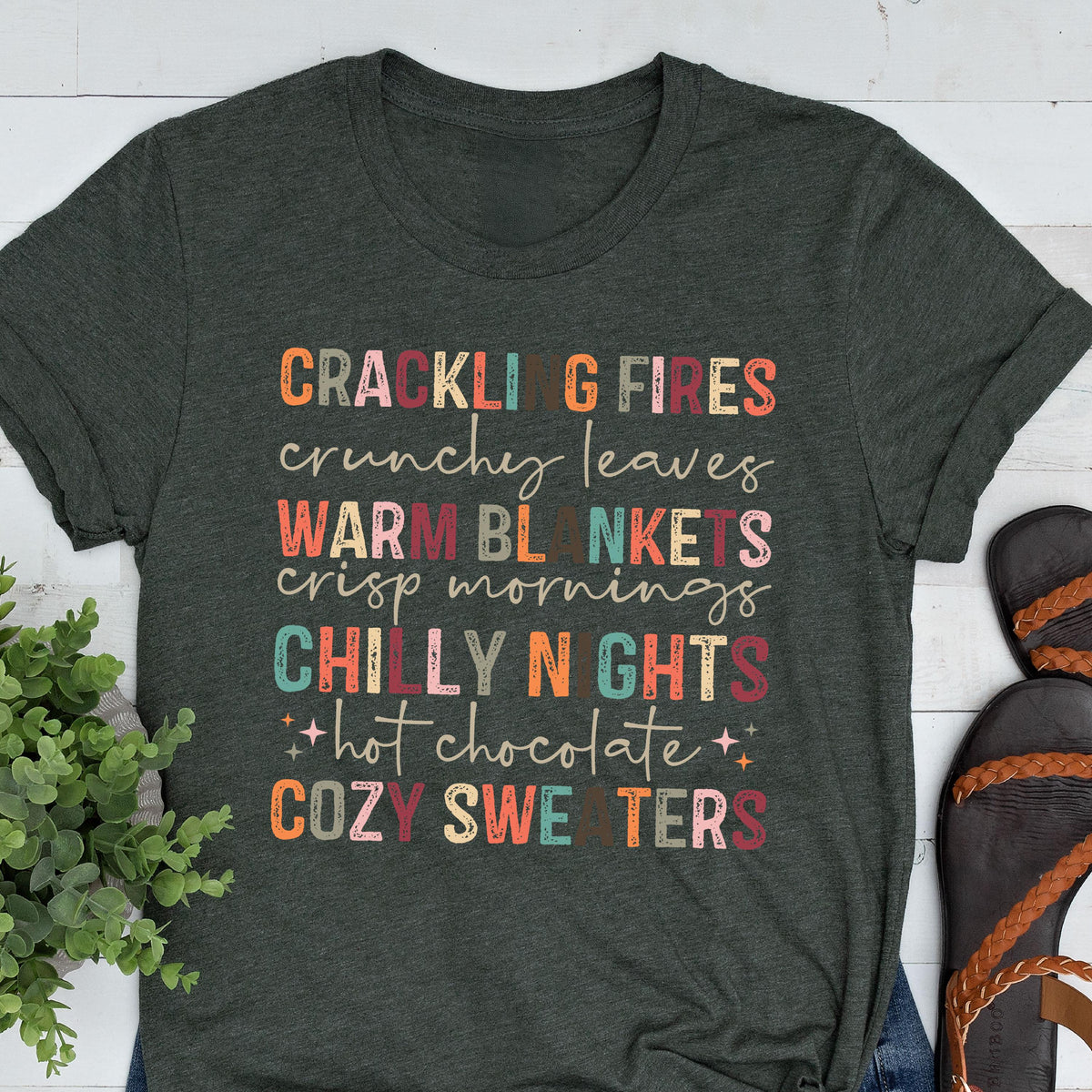 May include: Dark gray t-shirt with colorful text: "CRACKLING FIRES, crunchy leaves, WARM BLANKETS, crisp mornings, CHILLY NIGHTS, hot chocolate, COZY SWEATERS." The shirt has a crew neck and short sleeves.