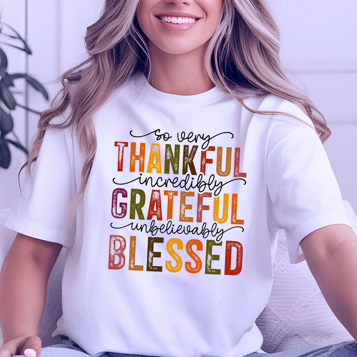 May include: A white short-sleeved t-shirt featuring the text "so very THANKFUL incredibly GRATEFUL unbelievably BLESSED" printed in a mix of black cursive script and block letters in shades of orange, yellow, brown, and dark green.